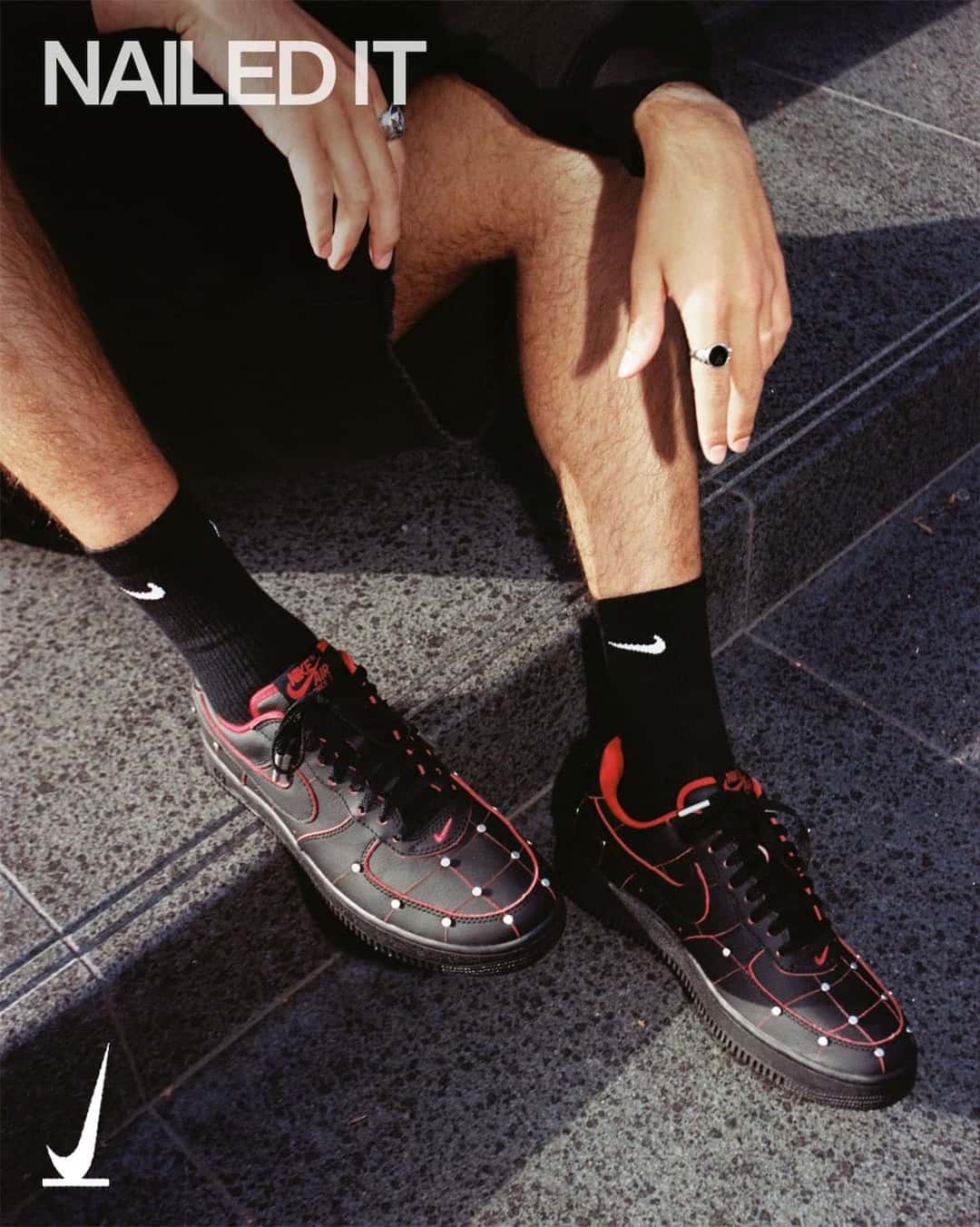 Persona sentada en una escalera con el Nike Air Force 1 '01 Nightmare puesto, en cuero negro con costuras rojas en cuadrícula y piezas metálicas plateadas que simulan clavos en la puntera, en la imagen oficial de SNKRS con la frase Nailed It