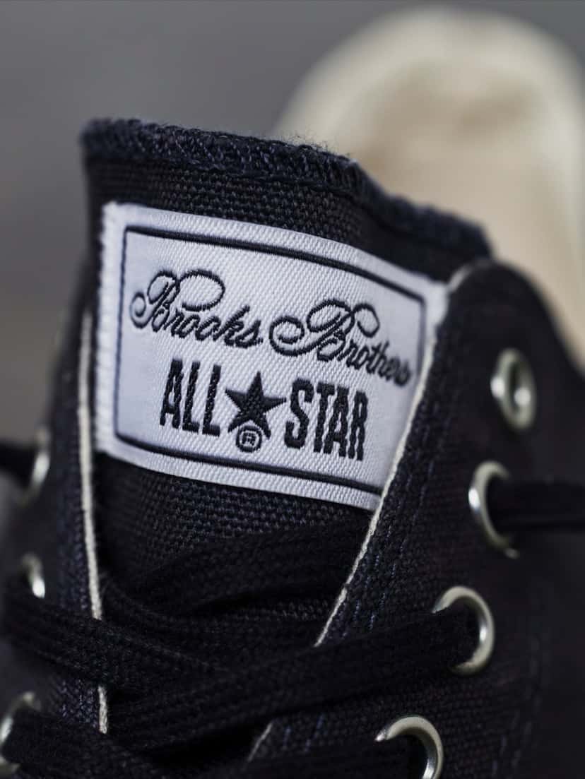 Etiqueta de la lengüeta de la versión azul marino, con la firma Brooks Brothers en cursiva sobre el logo ALL STAR con la estrella, bordados en azul sobre fondo blanco