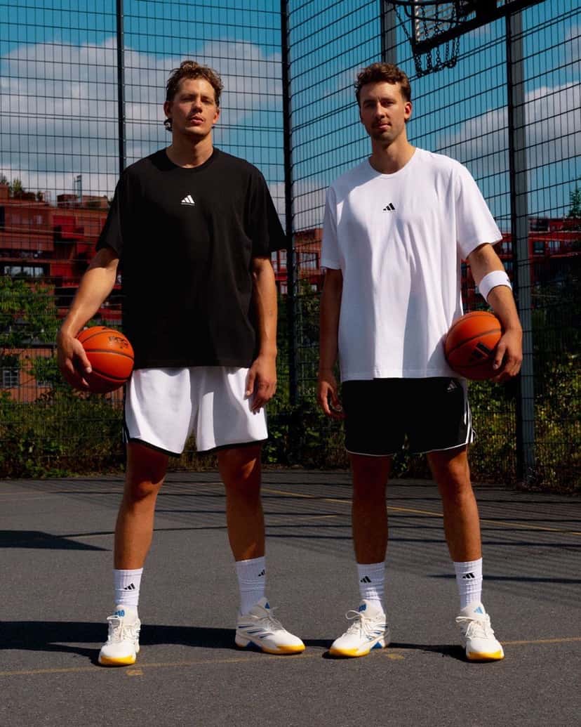 Franz y Moritz Wagner parados en una cancha de básquet al aire libre en Berlín, uno con remera negra y otro con remera blanca de adidas, cada uno con una pelota y las Crazy Energy blancas con suela amarilla puestas