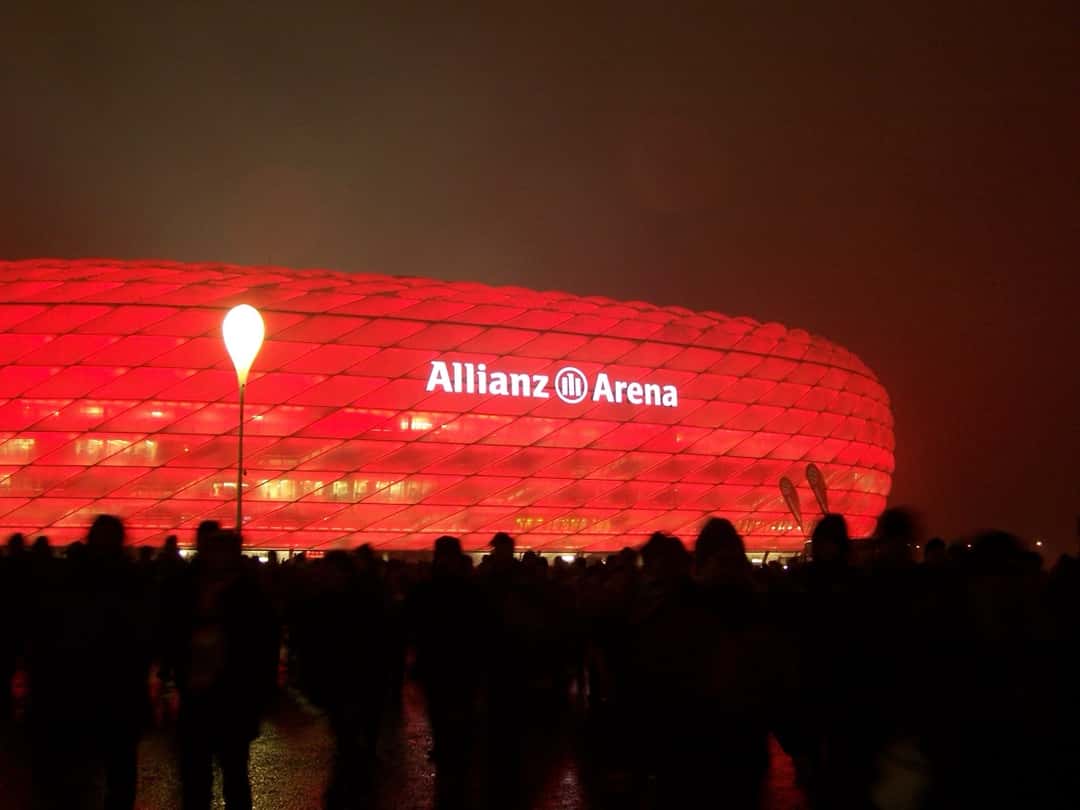 El Allianz Arena de Múnich iluminado de rojo de noche, con hinchas caminando hacia el estadio; foto de archivo de 2012 (foto: OdinFK, Wikimedia Commons, licencia CC BY-SA 3.0)
