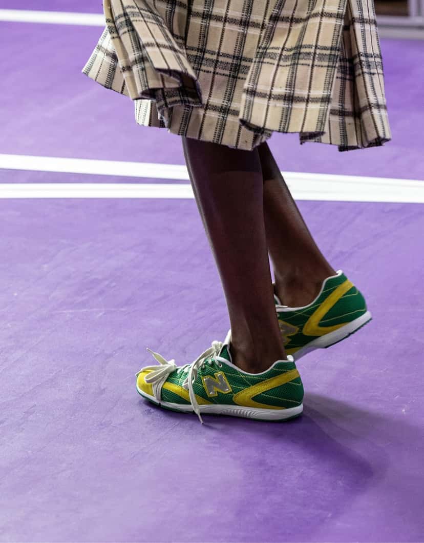 Foto oficial del desfile Primavera/Verano 2027 de Miu Miu: New Balance RC150 verdes con detalles amarillos y logo N plateado, con una pollera escocesa beige, sobre la pista violeta