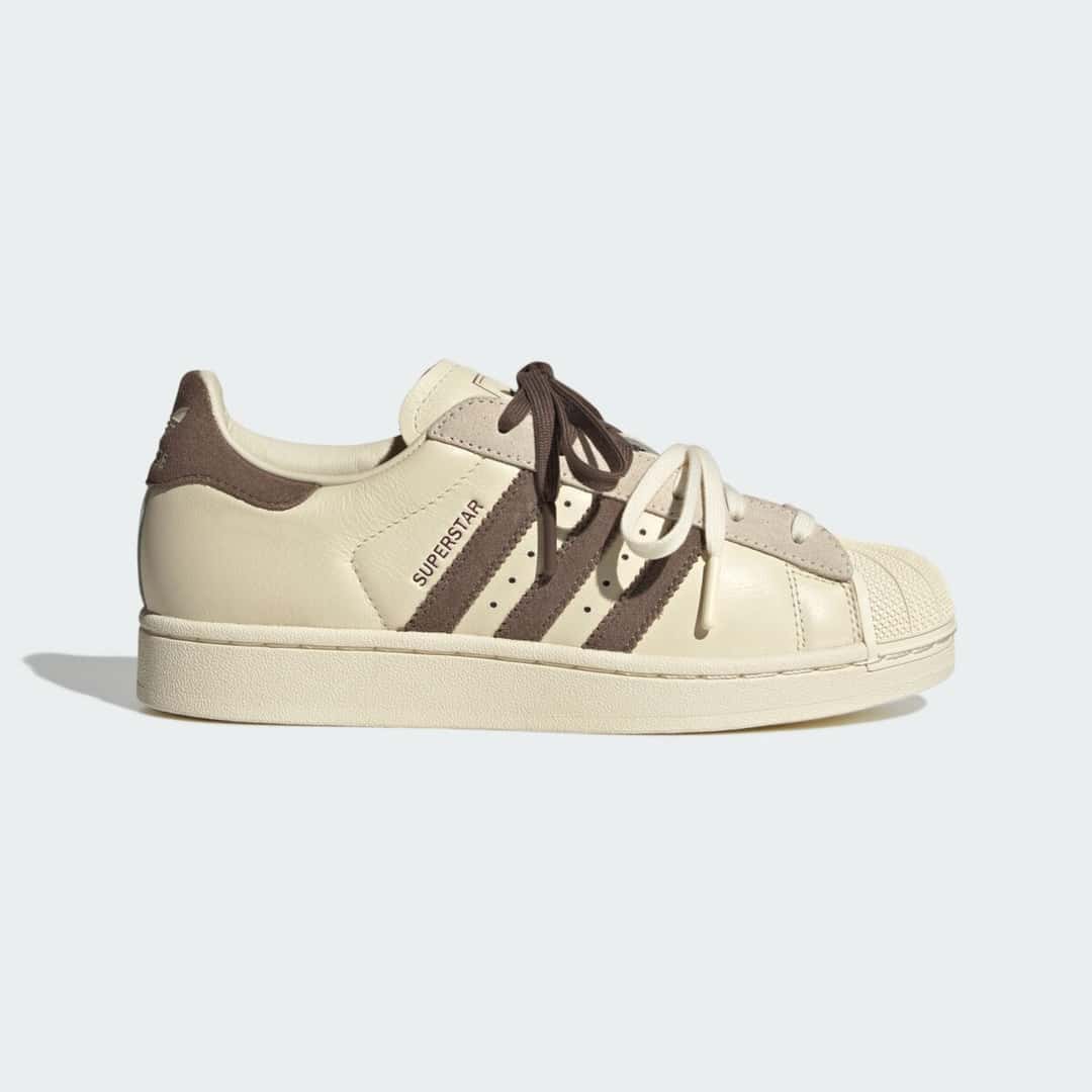 Molly-Mae x adidas Superstar Latte de perfil sobre fondo gris claro, con cuero color crema, Three Stripes marrones, doble cordón crema y marrón y la puntera de goma shell toe