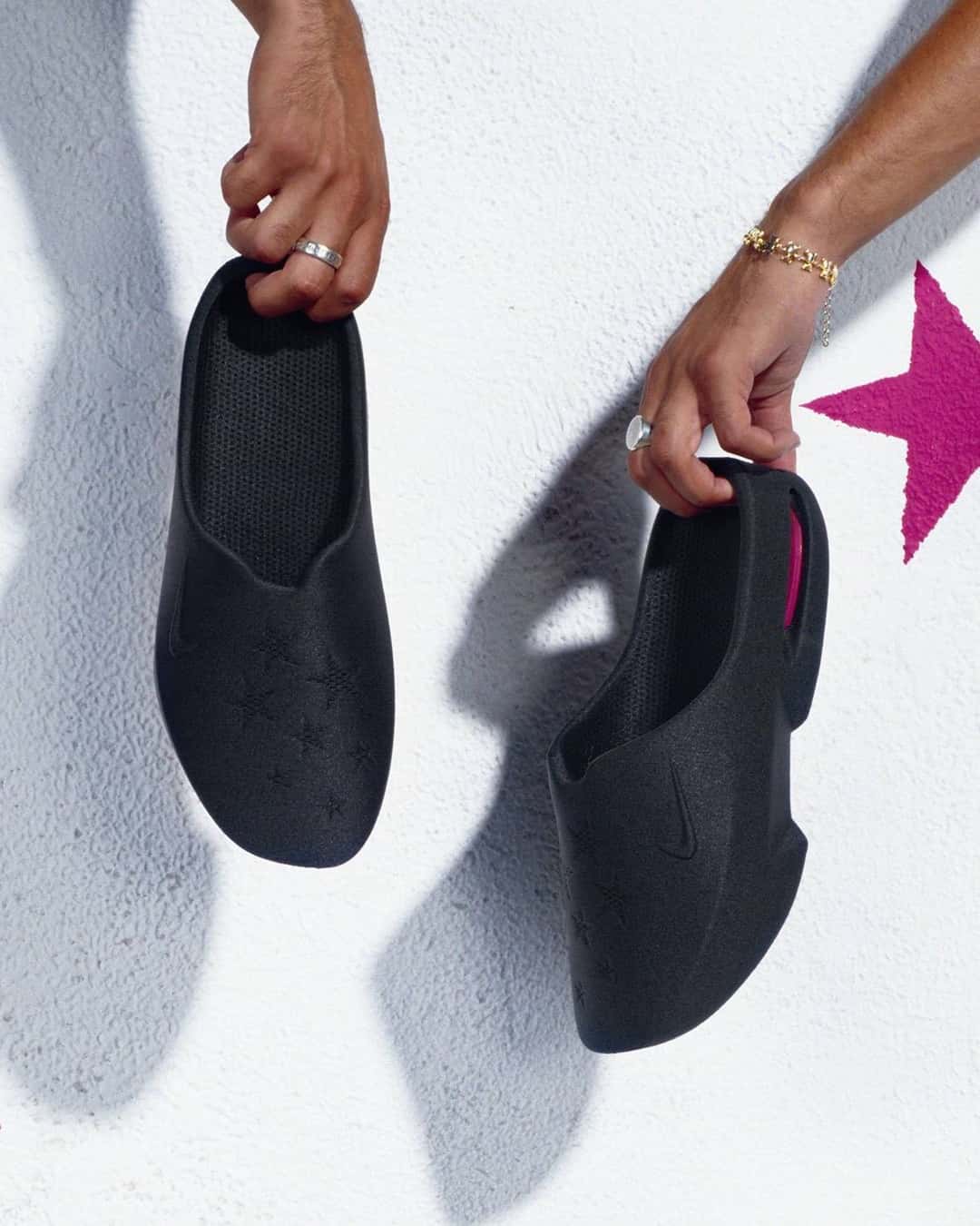 Dos manos sostienen las Nike Air Max Mule negras de Masyn contra una pared blanca, con las estrellas en relieve en la capellada y una estrella rosa pintada en la pared