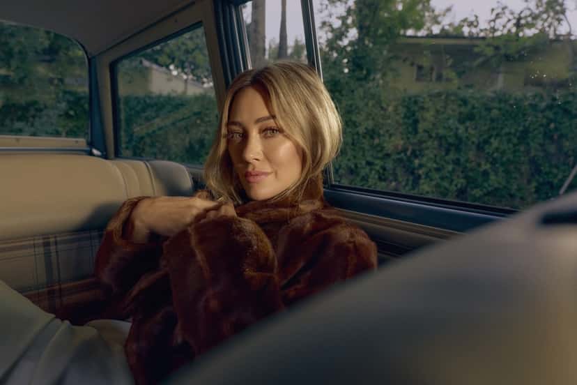 Hilary Duff sentada en el asiento trasero de un auto, con un abrigo de piel marrón y el pelo rubio suelto, en una foto de prensa de la etapa de 