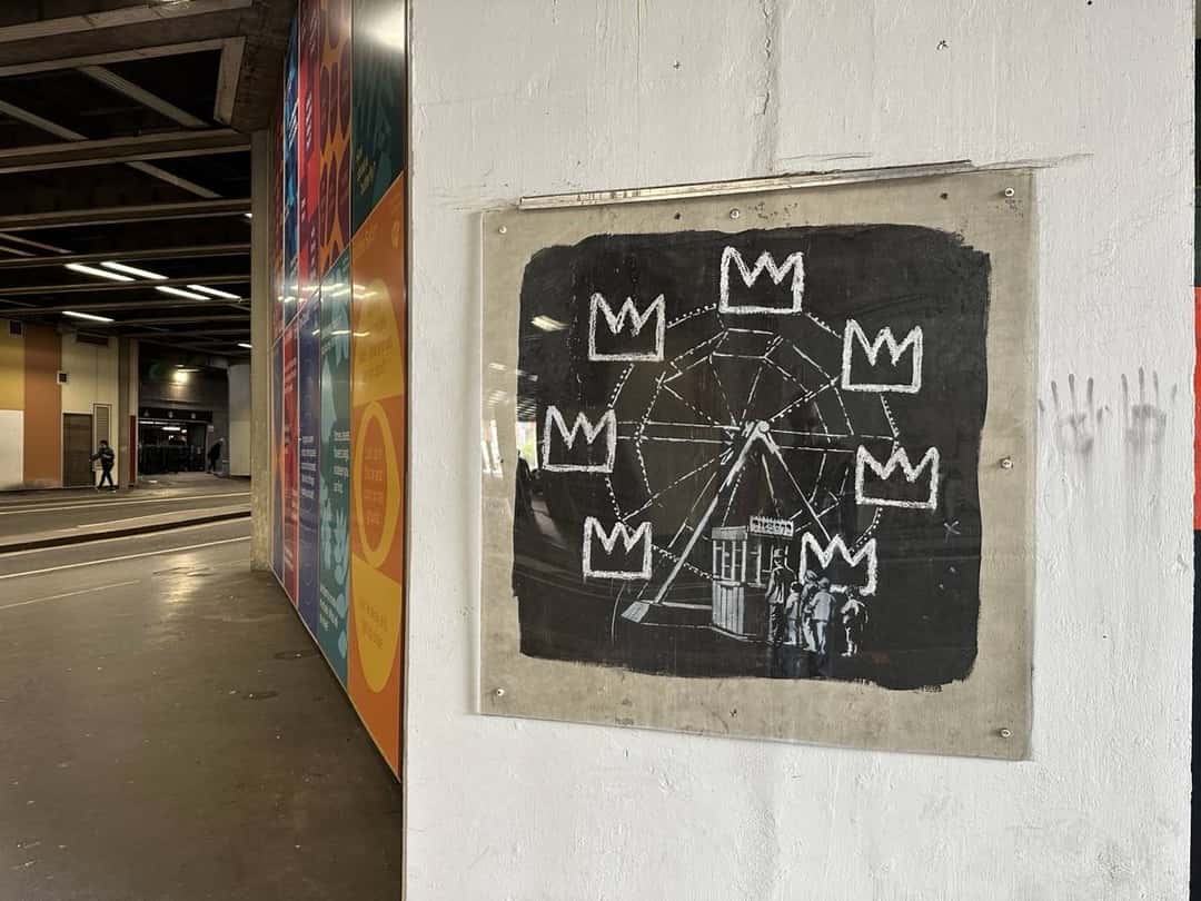 Un mural de Banksy en homenaje a Basquiat: una vuelta al mundo dibujada en blanco con coronas a su alrededor, sobre un fondo negro, protegido por un acrílico en una pared blanca de un pasaje (imagen que representa al artista). Foto: Matt Brown, CC BY 2.0, vía Wikimedia Commons