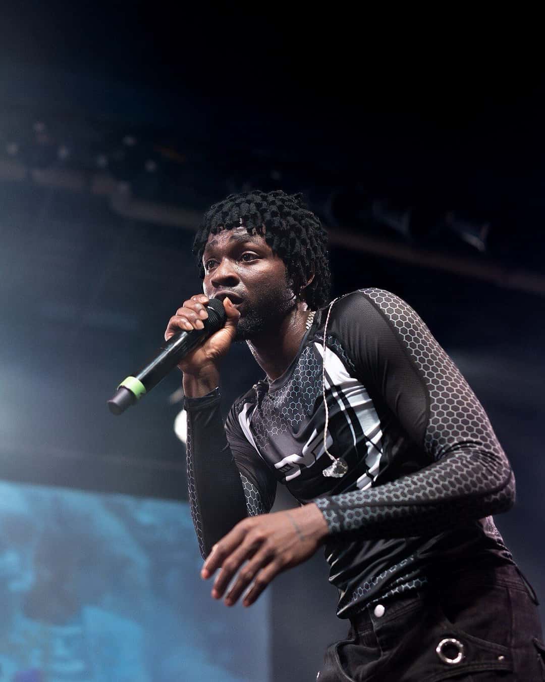 Foto de archivo de Omah Lay en vivo en 2023: canta con un micrófono en la mano, con una remera de mangas de red y estampa geométrica, bajo luces azules. Foto: Warner Records, Wikimedia Commons, CC BY-SA 4.0