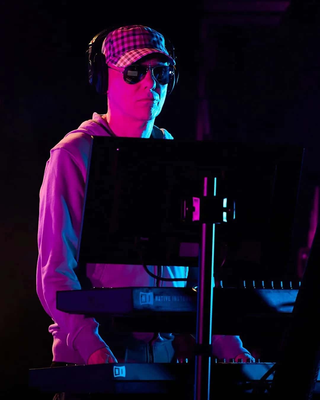 Chris Lowe tocando teclados bajo luces rosas y azules, con gorra a cuadros, anteojos oscuros, auriculares y buzo claro; foto publicada por Pet Shop Boys