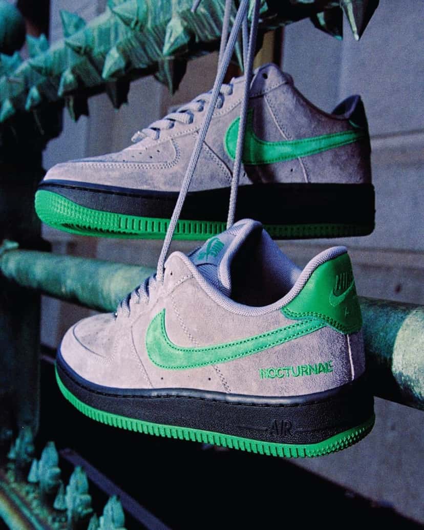 Foto de campaña de dos Nike SB Air Force 1 de Nocturnal colgadas de los cordones de una reja verde con puntas, con upper de gamuza gris, Swoosh y talón verdes, mediasuela negra, suela verde y la palabra NOCTURNAL bordada en verde en el lateral del talón