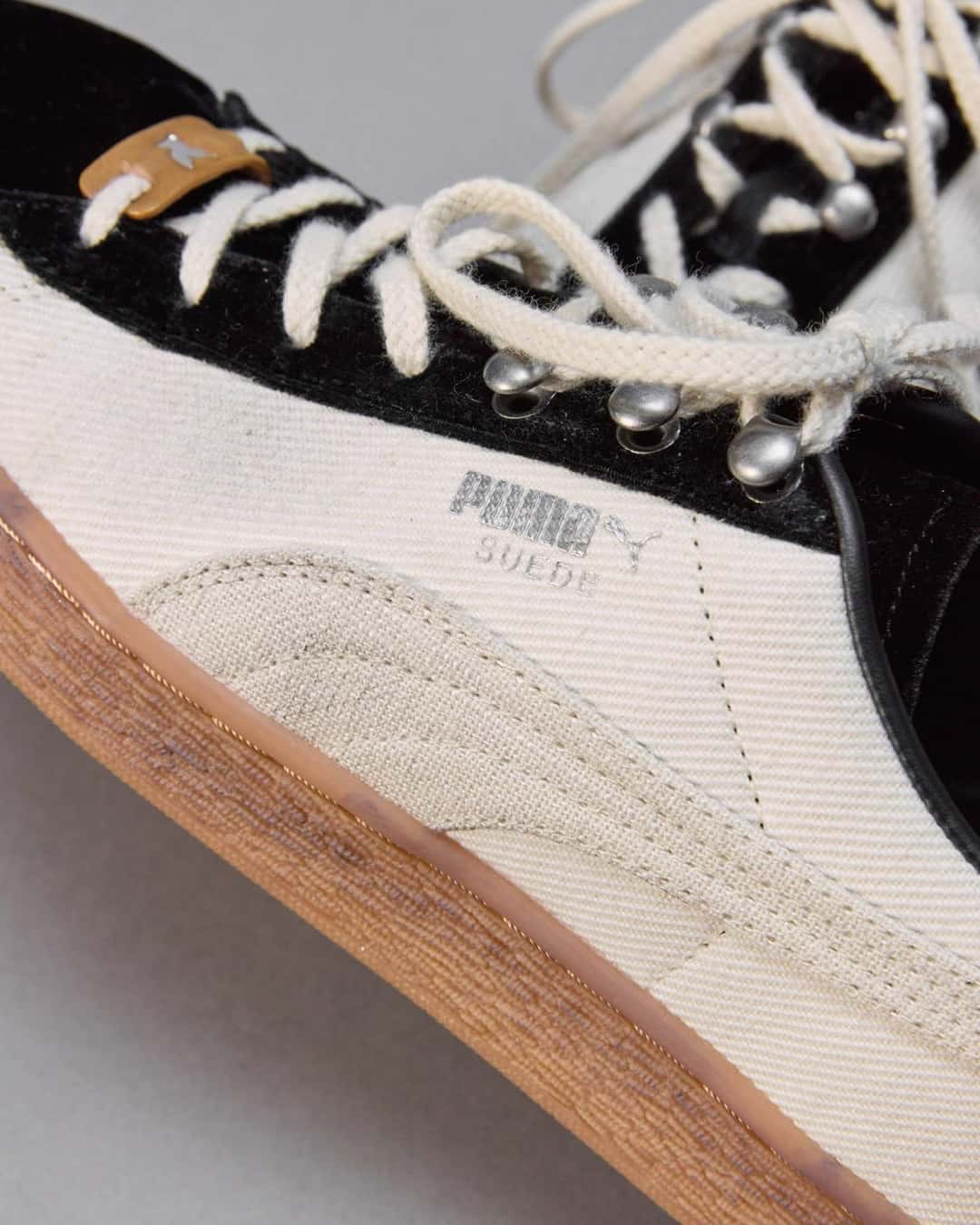 Detalle lateral del PUMA x MASU Curio Suede, con los ganchos metálicos plateados para los cordones, la zona de cordones en gamuza negra, el cuerpo marfil con la franja Formstrip del mismo tono, la leyenda PUMA Suede impresa y la suela color caramelo