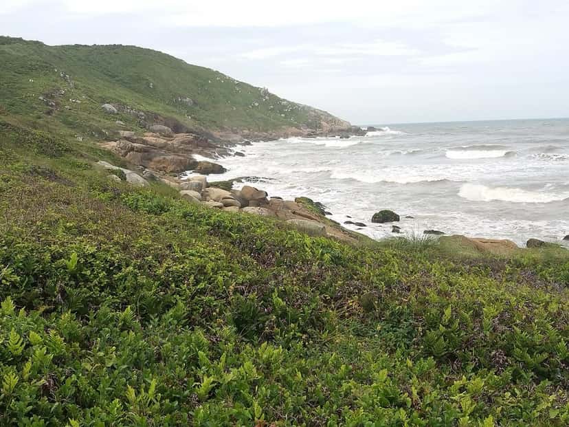 Costa rocosa de la Praia da Vila, en Imbituba, con olas que rompen junto a un morro cubierto de vegetación