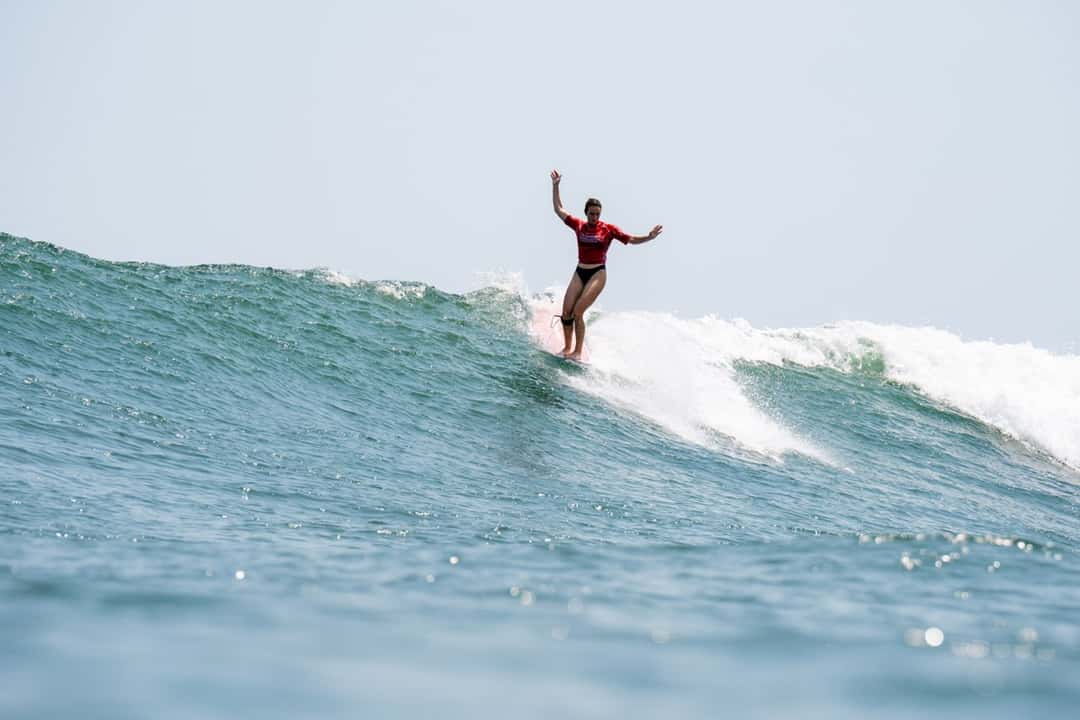 Rachael Tilly, de lycra roja, camina sobre el longboard con los brazos abiertos en el labio de una ola en El Sunzal