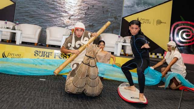 Un surfista con traje de neoprene hace la postura de surf sobre una tabla en el escenario, junto a un actor con un caballito de totora y bailarines con telas celestes que simulan el mar