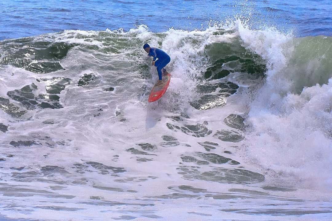 Un surfista con traje azul y tabla roja baja una ola grande que rompe en espuma en Punta Rocas, Perú