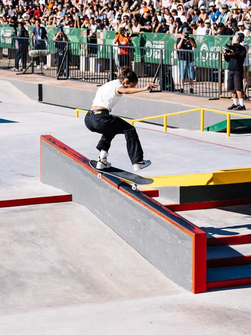Rayssa Leal, con remera blanca y pantalón negro, se desliza sobre el borde de un cajón gris de la pista de street de Luque, con el público detrás de las vallas
