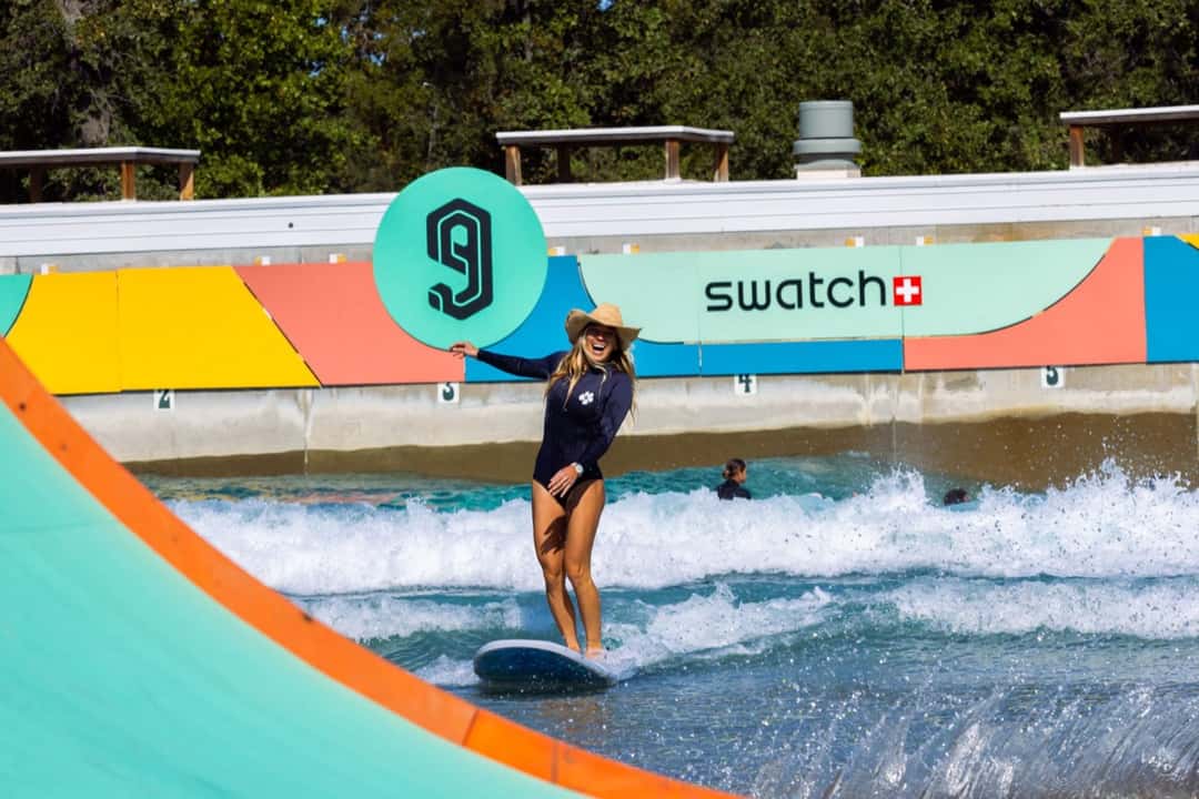 Coco Ho, con traje negro y sombrero de ala ancha, saluda mientras surfea en Waco Surf frente al cartel de Swatch y el logo de los Nines, en la edición 2024