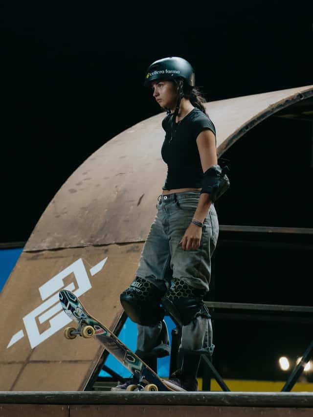 Una skater con casco negro y rodilleras espera parada en el borde de la rampa vertical con la tabla lista para tirarse, de noche, en una fecha anterior del Vert Battle (foto de archivo)
