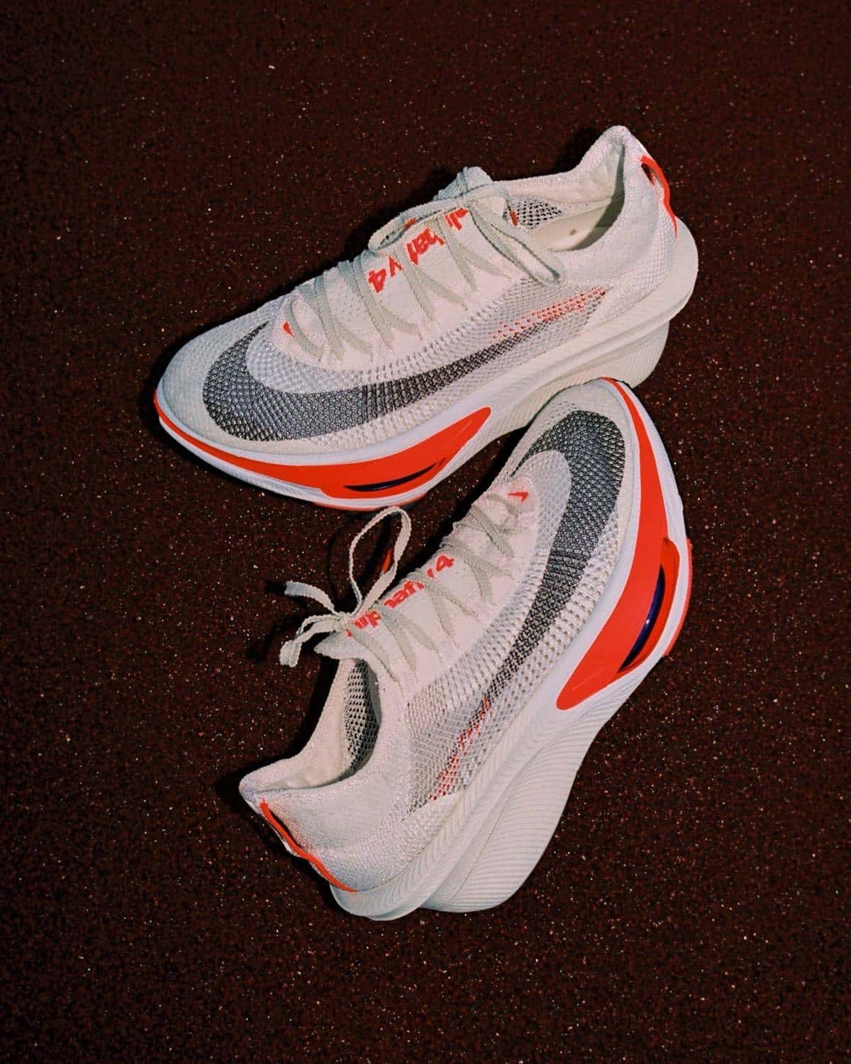 Par de Nike Alphafly 4 blancas con Swoosh gris oscuro y detalles rojos, una sobre otra sobre un fondo de tierra rojiza