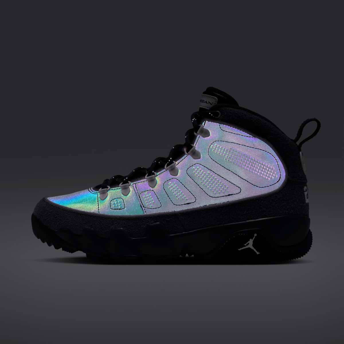 Air Jordan 9 Retro Boot NRG Anthracite de perfil sobre fondo gris oscuro, con los paneles laterales reflectivos brillando en tonos lila, celeste y verde, gamuza gris oscura en la base y el talón y suela negra con el Jumpman blanco