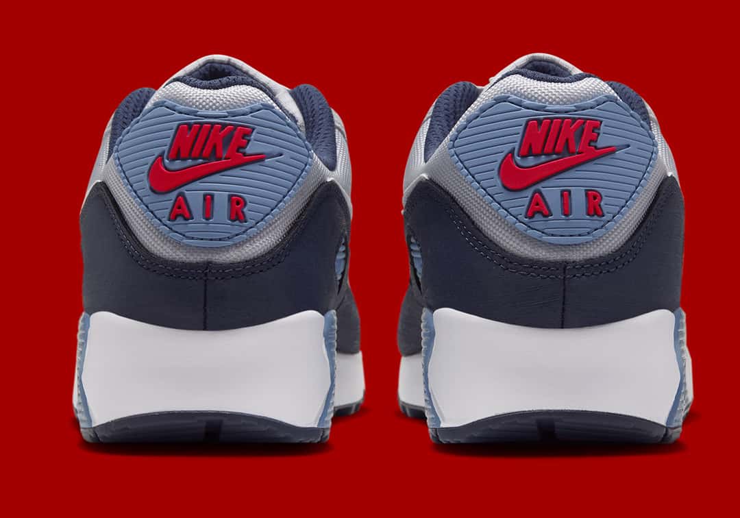 Talones del Nike Air Max 90 Geyser Grey Work Blue sobre fondo rojo, con el parche Work Blue con el logo Nike Air rojo, la base azul marino y la mediasuela blanca