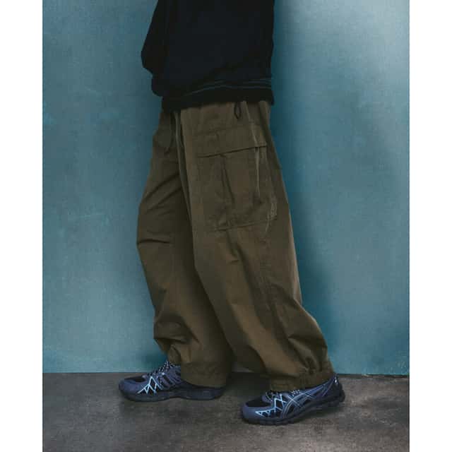 Foto de campaña de la ASICS Utility Collection con el GEL-KAYANO 20 en azul marino oscuro con la jaula de TPU en celeste, usado con un pantalón cargo verde oliva frente a una pared azul
