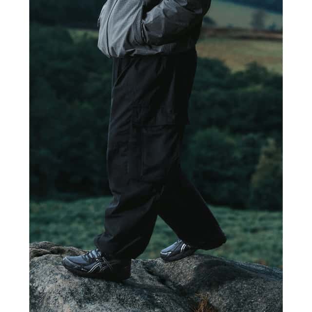 Foto de campaña de la ASICS Utility Collection con el GEL-VENTX GTX gris y negro con las rayas ASICS plateadas, usado con pantalón cargo negro sobre una roca en un paisaje de campo