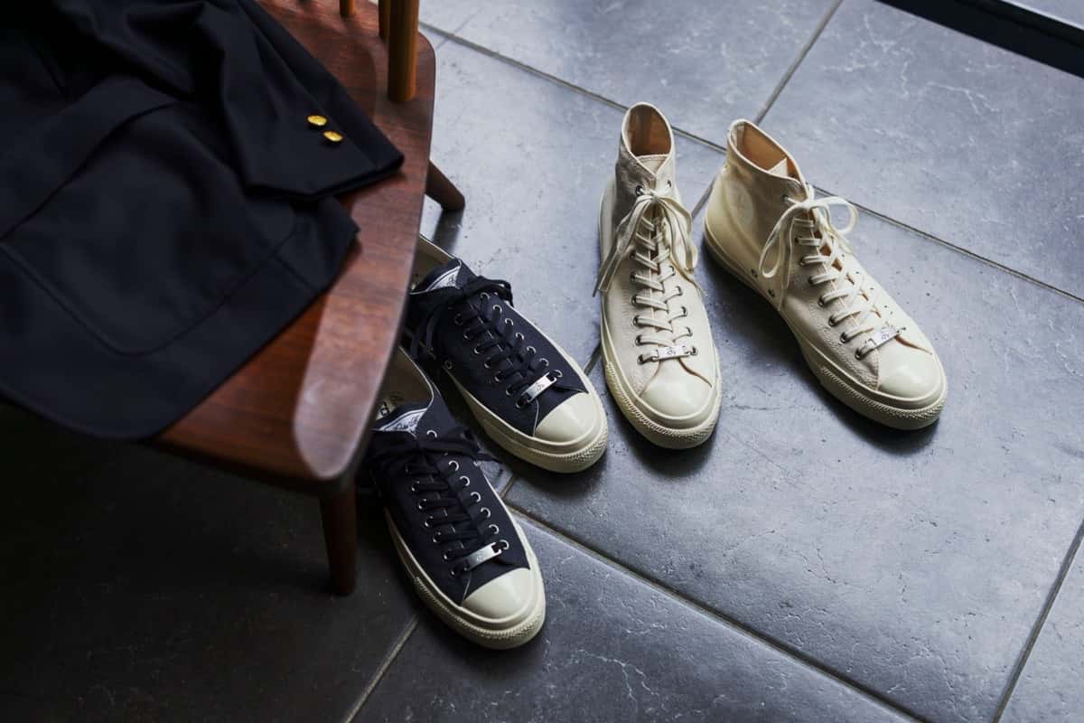 Dos pares de Brooks Brothers x Converse All Star Aged sobre un piso de baldosas gris oscuro: a la izquierda las Ox azul marino junto a una silla de madera con un saco azul con botones dorados, a la derecha las Hi color crudo con cordones claros
