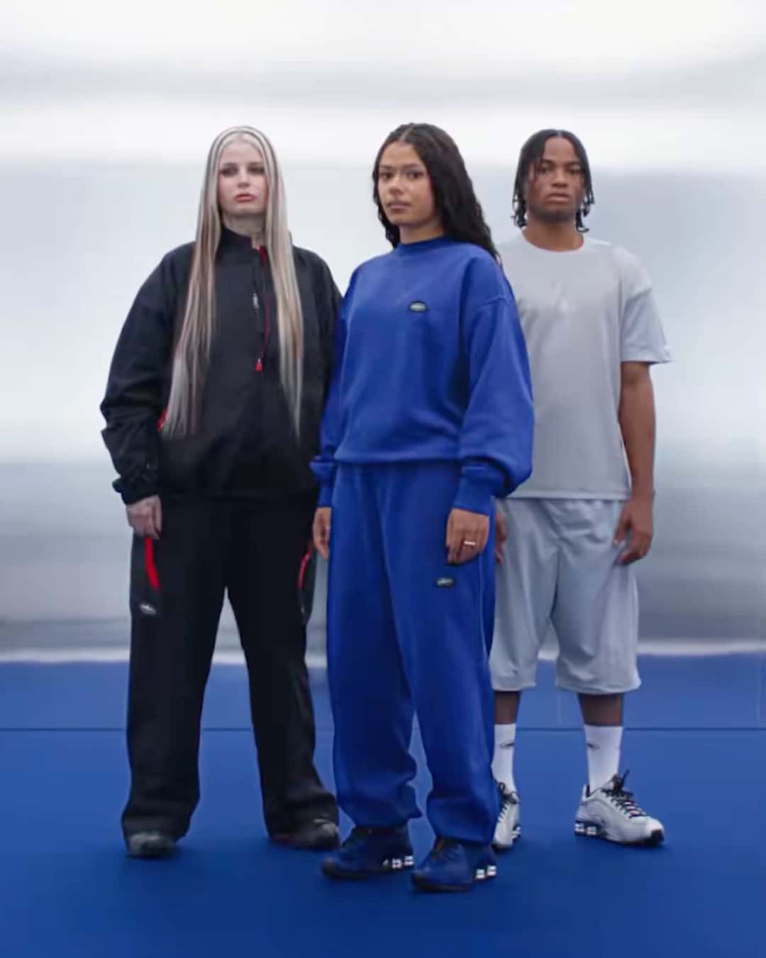 Tres modelos de la campaña de Corteiz x Nike de pie sobre un piso azul: una con conjunto negro, otra con conjunto azul y un chico con remera gris y shorts, cada uno con un color distinto del Shox R419