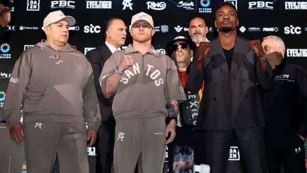 Canelo Álvarez, de buzo gris con la palabra «Santos», y Christian Mbilli, de traje oscuro, posan con los puños cerrados frente a un panel de sponsors en la conferencia de prensa de Riad