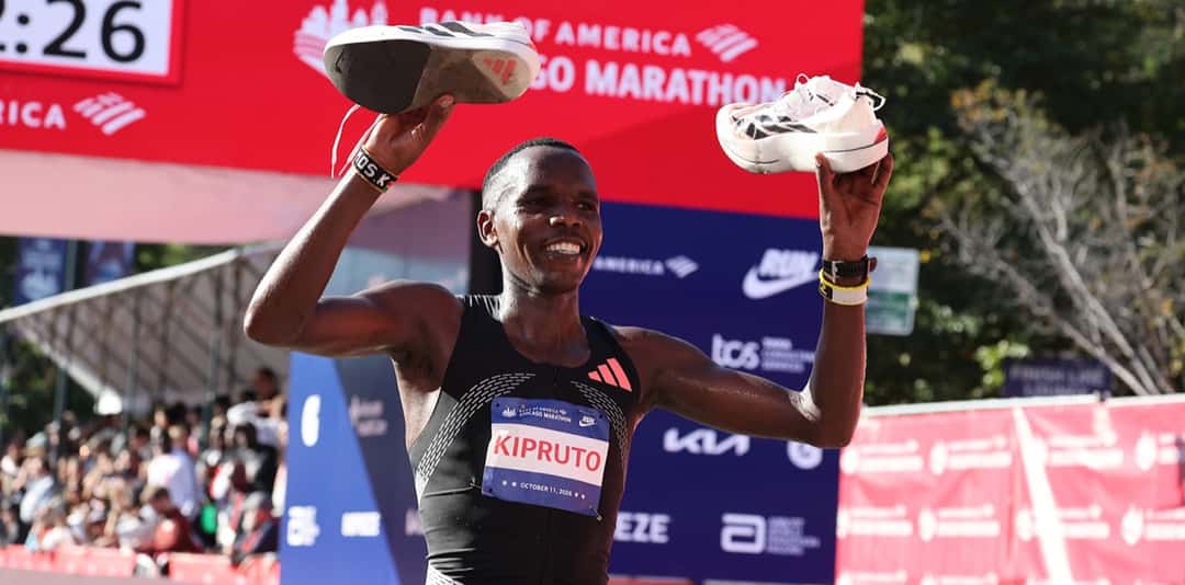 Amos Kipruto, con la musculosa negra y el dorsal con su apellido, sonríe y levanta un par de zapatillas en cada mano frente al cartel del Maratón de Chicago