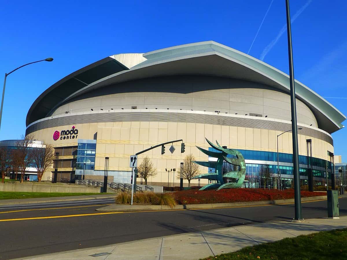 Imagen del estadio: el Moda Center de Portland, un estadio redondo de techo plateado, con una escultura verde en la vereda y el cielo azul