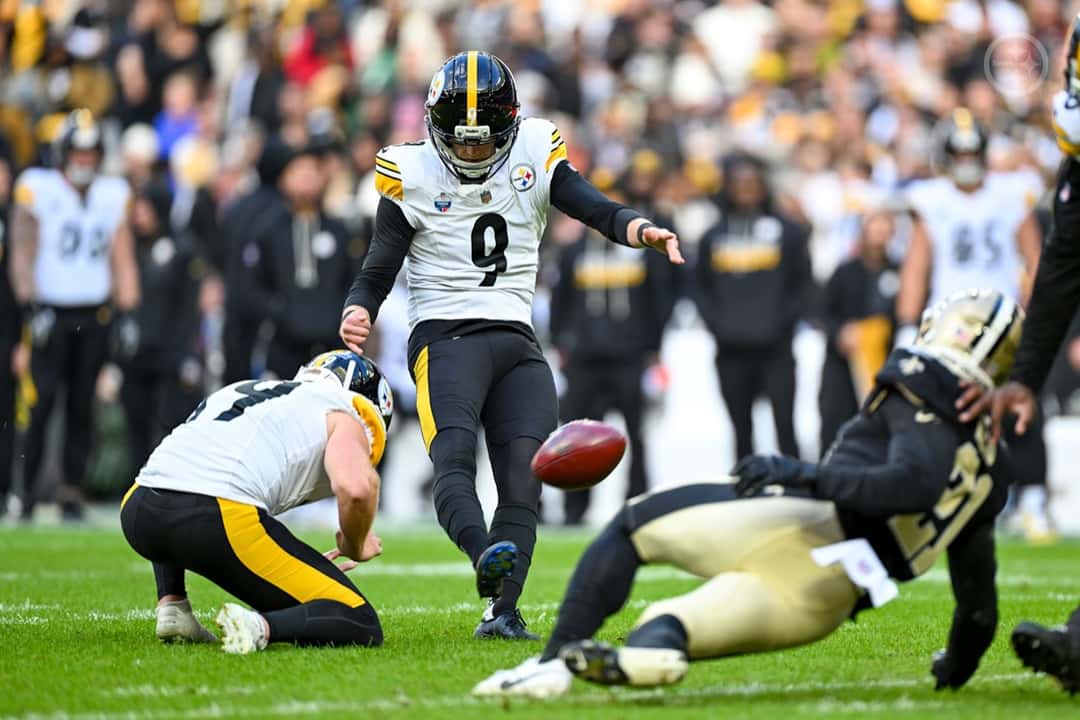 Chris Boswell, con la camiseta blanca número 9 de los Steelers, patea un gol de campo mientras un compañero sostiene la pelota y un jugador de los Saints se tira a bloquear