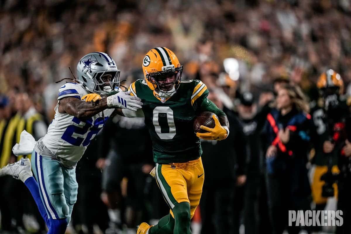 Matthew Golden, de los Green Bay Packers, con la camiseta verde número 0, corre con la pelota bajo el brazo derecho mientras un defensor de los Dallas Cowboys, de blanco con el número 26, intenta frenarlo, con la tribuna de Lambeau Field detrás