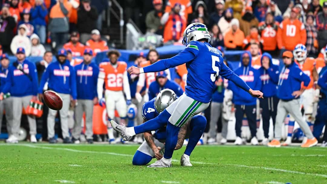 Jason Myers, pateador de los Seahawks con la camiseta azul número 5, le pega a la pelota sostenida por un compañero, con la tribuna naranja de los Broncos de fondo