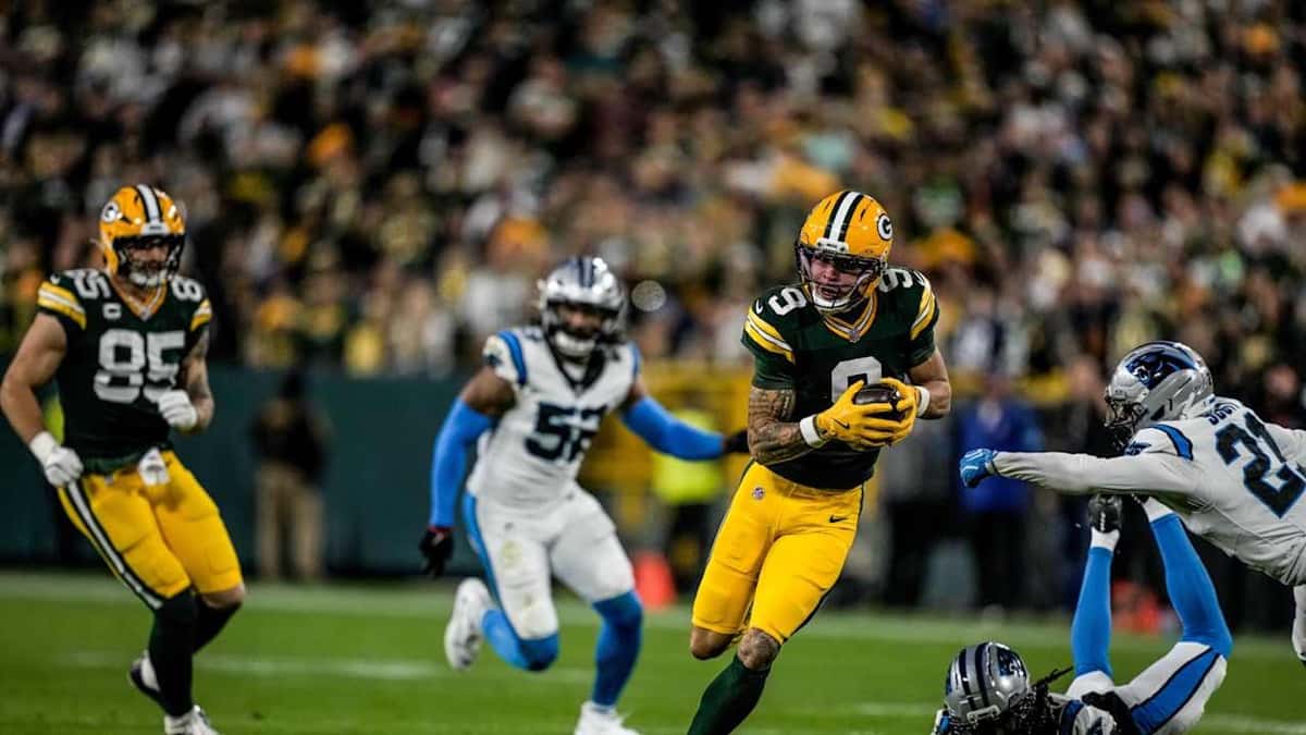 Christian Watson, de los Packers, con la camiseta verde número 9, corre con la pelota mientras dos defensores de los Panthers, de blanco, intentan alcanzarlo en el Lambeau Field