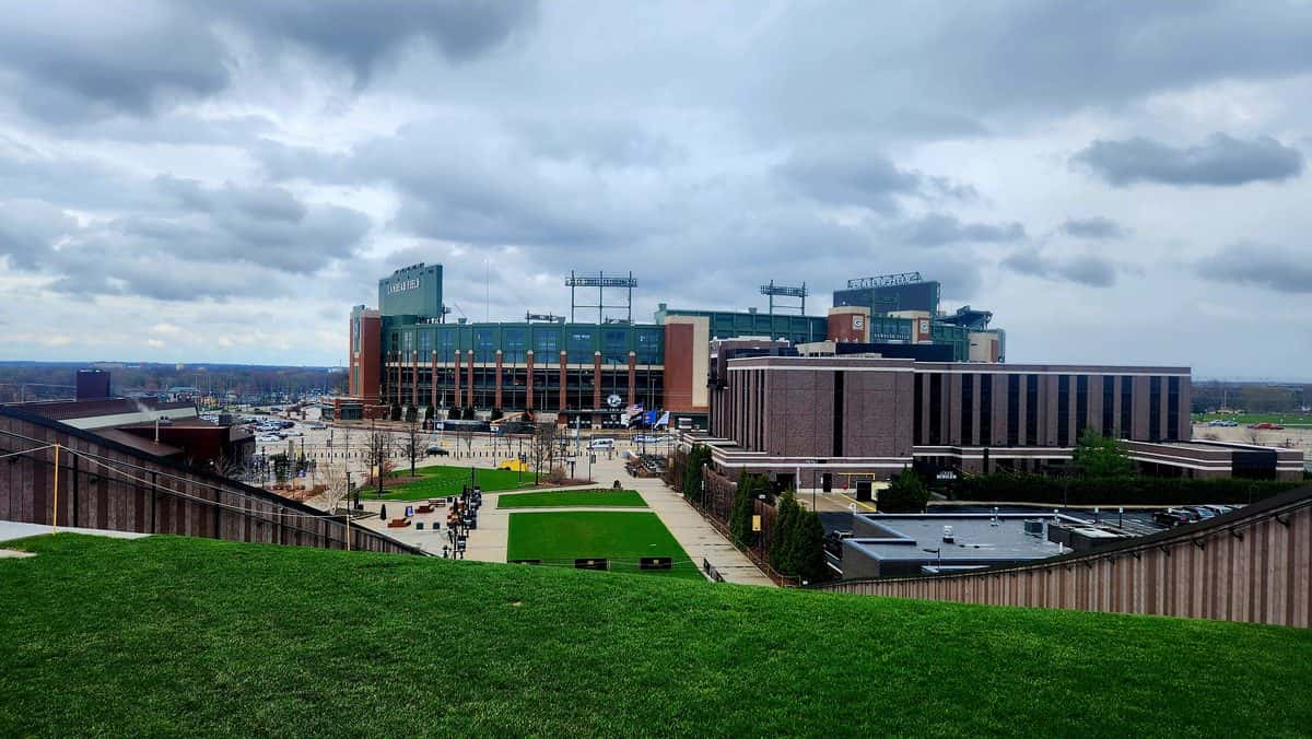 Imagen del estadio: el Lambeau Field de Green Bay visto a lo lejos desde una terraza con césped, bajo un cielo nublado