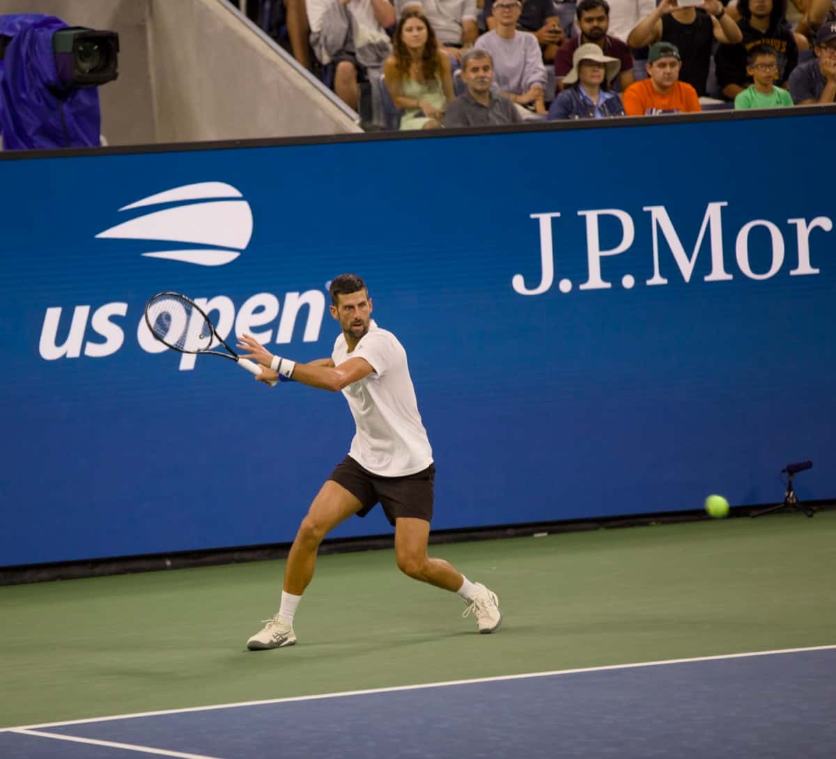 Foto de archivo: Novak Djokovic, de remera blanca y short negro, pega un revés en una cancha dura del US Open 2026, con el cartel del torneo detrás