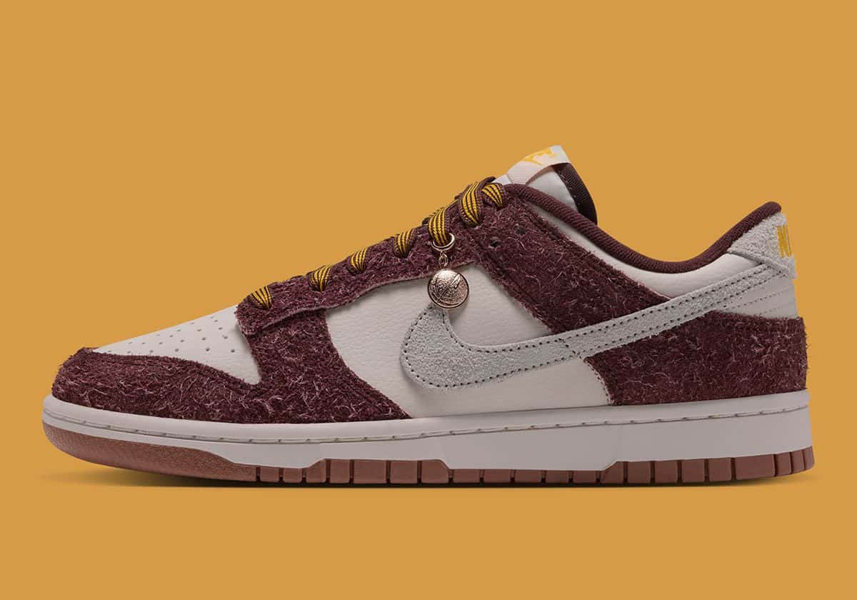 Nike Dunk Low Basketball Charm de perfil, con gamuza peluda bordó, base crema, Swoosh de gamuza crema, un dije metálico en los cordones y suela de goma marrón, sobre fondo mostaza