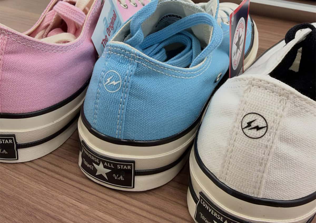 Talones de tres Converse Chuck Taylor bajas de fragment design en rosa, celeste y blanco sobre una mesa de madera, con el rayo de fragment y el parche Converse All Star