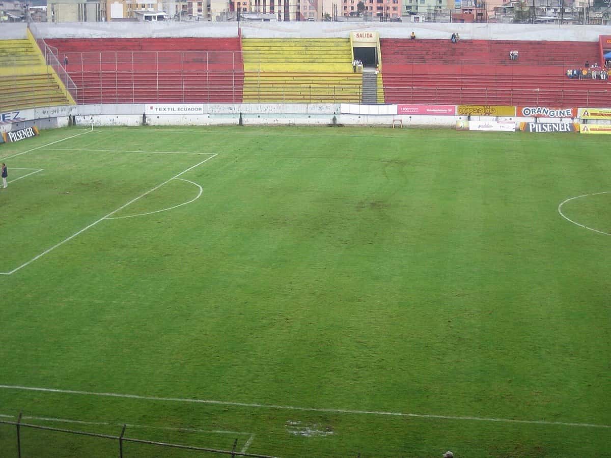 El estadio Gonzalo Pozo Ripalda, de Aucas, en el barrio de Chillogallo, en Quito, con las tribunas rojas y amarillas vacías y la cancha; foto de archivo de 2009 (foto: Albert7384, Wikimedia Commons, licencia CC BY-SA 4.0)