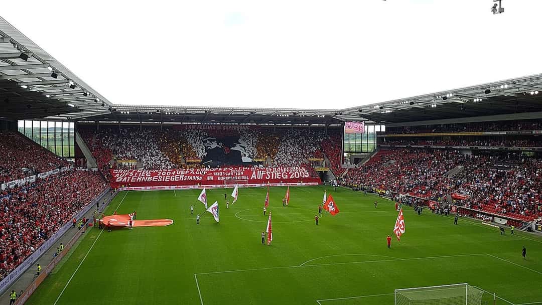 Las tribunas del estadio del Mainz llenas de hinchas, con banderas rojas y blancas en la cancha antes de un partido; foto de archivo de mayo de 2019 (foto: Lacu Schienred, Wikimedia Commons, licencia CC BY-SA 4.0)