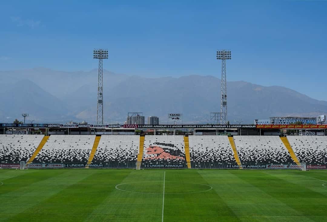 El estadio Monumental David Arellano, de Colo-Colo, en Santiago, con las tribunas blancas y negras vacías, la cancha y la cordillera de fondo; foto de archivo de diciembre de 2016 (foto: Carlos Figueroa, Wikimedia Commons, licencia CC BY-SA 4.0)