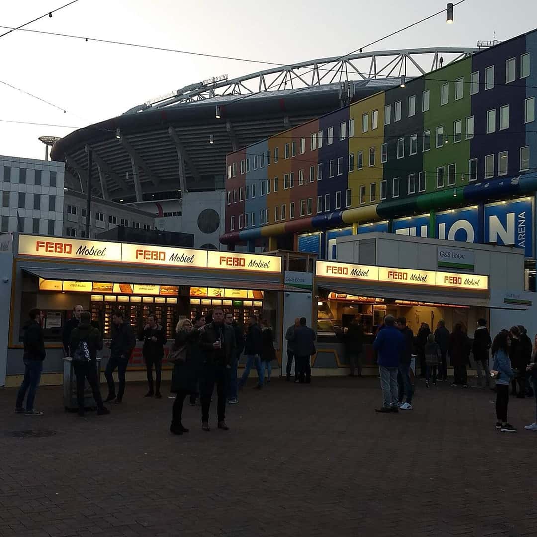 El Johan Cruyff Arena de Ámsterdam al atardecer, con su fachada de colores detrás de los puestos de comida de la explanada; foto de archivo de 2019 (foto: Edoderoo, Wikimedia Commons, licencia CC BY-SA 4.0)