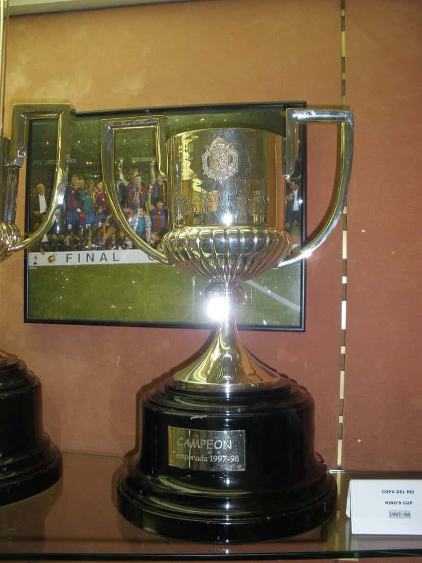 El trofeo de la Copa del Rey, una copa plateada con dos asas sobre una base negra, exhibido en una vitrina con la placa de campeón de la temporada 1997-98 (foto de archivo: Ignasi, Wikimedia Commons, CC BY-SA 4.0)