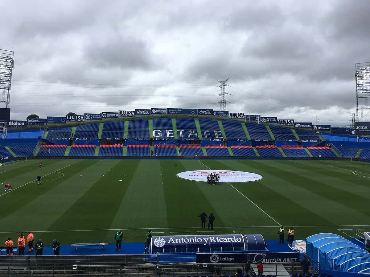 El Coliseum, estadio del Getafe, con la cancha rayada, las tribunas azules con la palabra GETAFE y los jugadores en la entrada en calor bajo un cielo nublado; foto de archivo de abril de 2018 (foto: Vincenzo.togni, Wikimedia Commons, licencia CC BY-SA 4.0)
