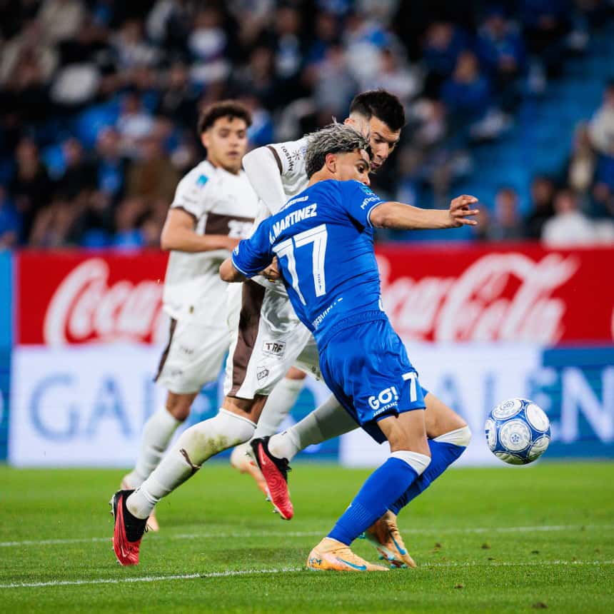 Ronaldo Martínez, con la camiseta azul número 77 de Vélez, protege la pelota de espaldas a un defensor de Platense, de camiseta blanca, en el estadio José Amalfitani el lunes 5 de octubre (foto: Hernán Mauricio, prensa oficial de Vélez)