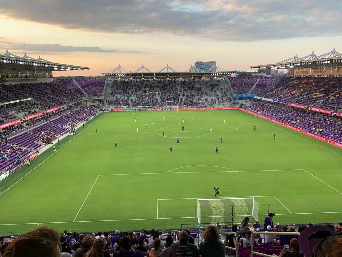 La cancha y las tribunas violetas del estadio de Orlando City durante un partido, vistas desde lo alto de una platea al atardecer; foto de archivo de 2021 (foto: timoteeburns, Wikimedia Commons, licencia CC BY-SA 3.0)