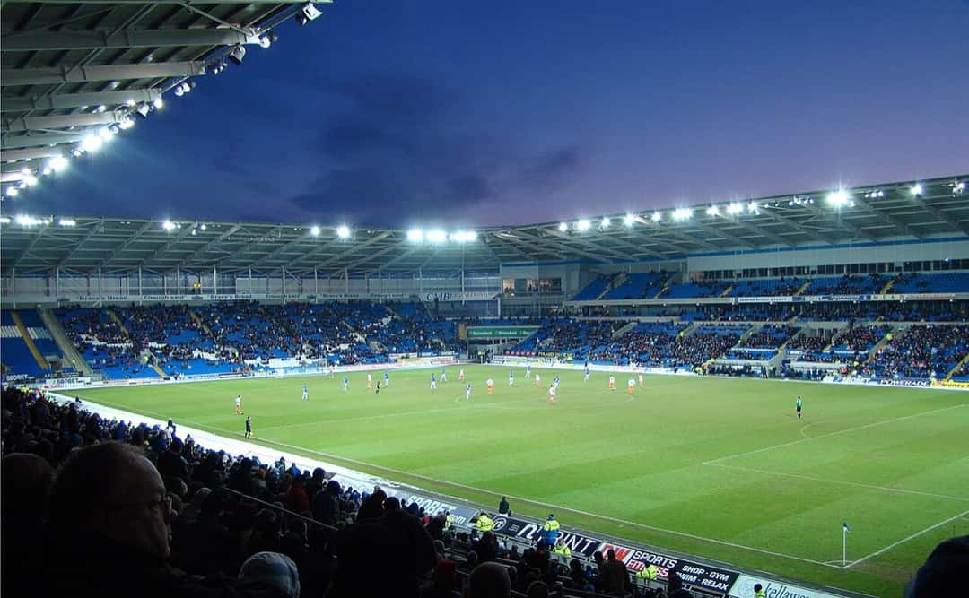 El Cardiff City Stadium iluminado de noche durante un partido, con las tribunas azules y el campo de juego (foto de archivo: Jon Candy, Wikimedia Commons, CC BY-SA 2.0)