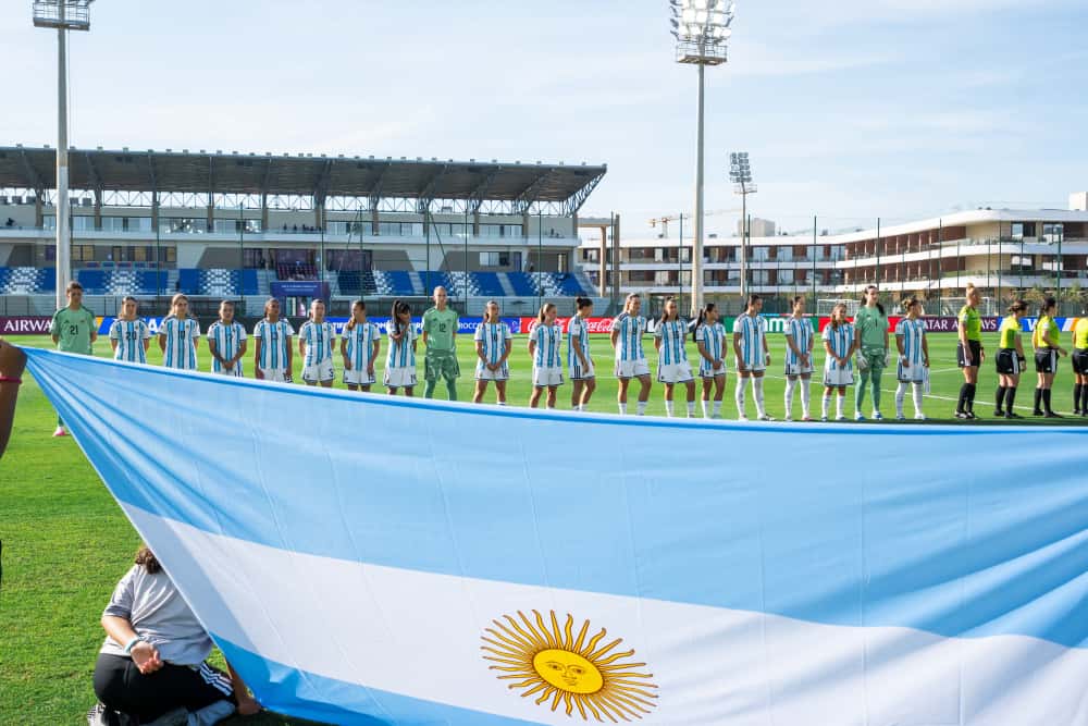 Las jugadoras de la Selección argentina femenina Sub-17, con camiseta celeste y blanca, formadas para los himnos detrás de una bandera argentina gigante en el Complejo Mohammed VI de Rabat; foto de archivo del Mundial (foto: prensa AFA)