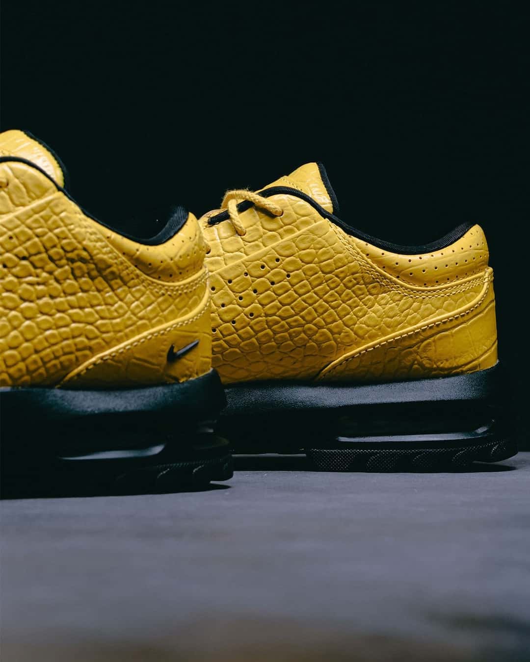 Vista lateral cercana del Nike Air Max Goadome Low Yellow Croc, con el cuero amarillo grabado tipo cocodrilo, cordones amarillos, swoosh negro chico y la base negra con la cámara de aire visible, sobre fondo negro