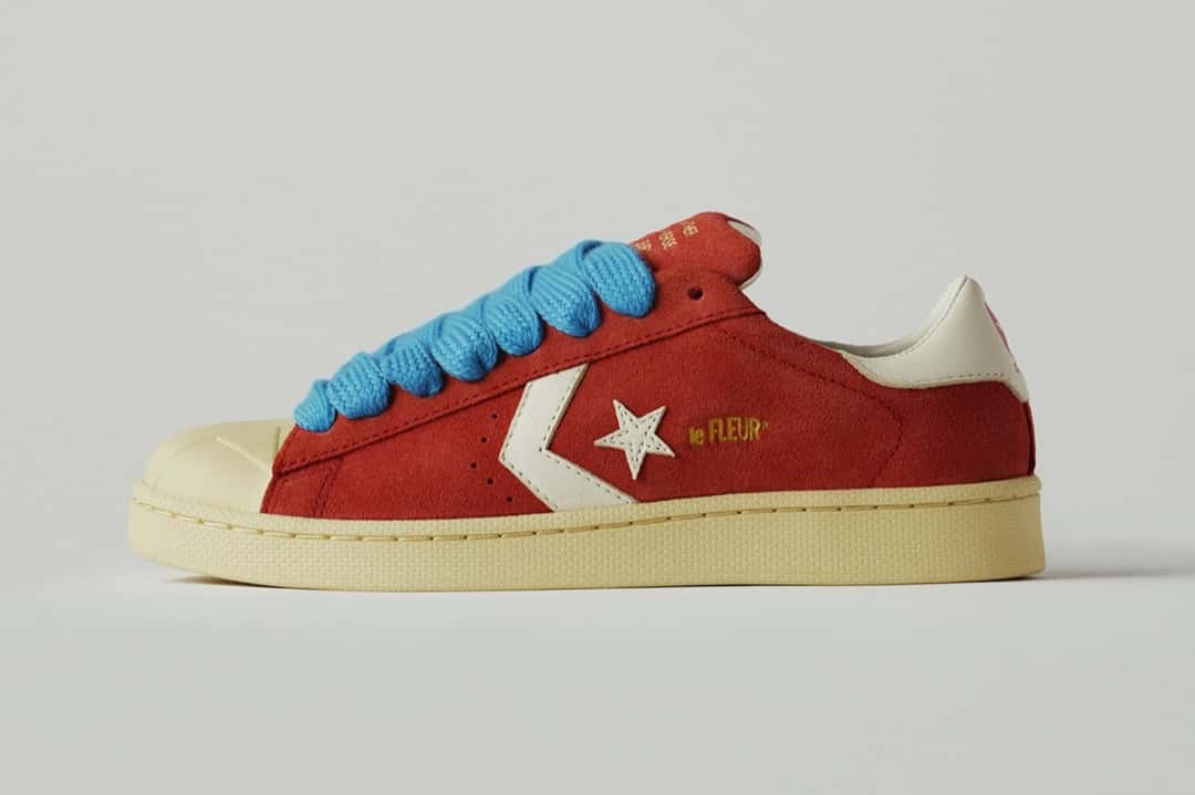 Converse Pro Leather Molten Lava de GOLF le FLEUR* de perfil sobre fondo gris claro, con upper de gamuza roja, la estrella y el chevron blancos, cordones gruesos celestes, puntera de goma color crudo y mediasuela crema
